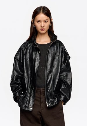 Młoda kobieta z długimi ciemnymi włosami, ubrana w błyszczącą czarną, oversized skórzaną kurtkę, czarną prążkowaną bluzkę i ciemnobrązowe spodnie, z rękami w kieszeniach.