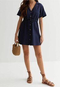 Marinblå klänning med korta ärmar, insydd midja och en framficka. Kombinerad med bruna sandaletter med remmar och en vävd handväska.