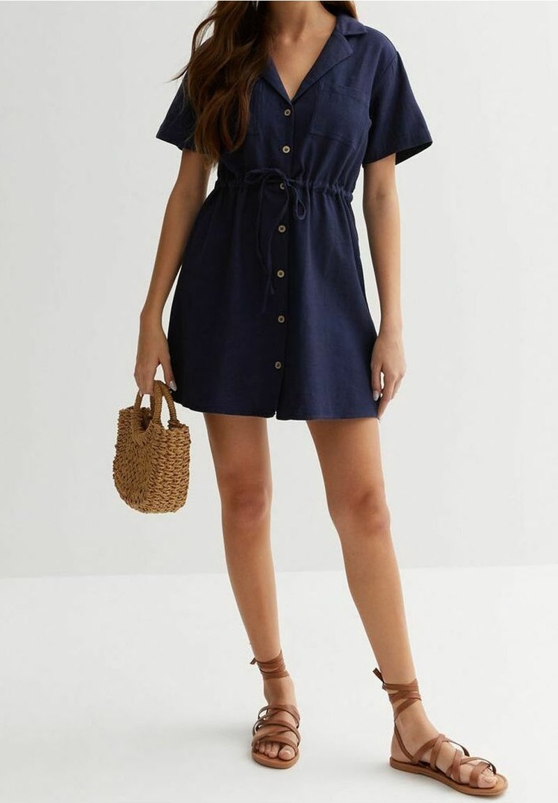 Marinblå klänning med korta ärmar, insydd midja och en framficka. Kombinerad med bruna sandaletter med remmar och en vävd handväska.