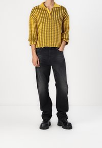 Żółty sweter z pionowymi paskami, kołnierzem na trzy guziki i rękawami do łokcia, zestawiony z ciemnymi jeansami i czarnym obuwiem.