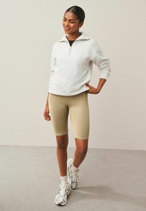 Giovane donna con pantaloncini da ciclismo beige, felpa bianca con zip, calzini bianchi e scarpe da ginnastica, in piedi con una mano sul fianco contro uno sfondo neutro.