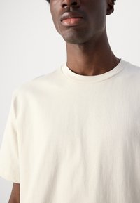 Tricou din bumbac bej, cu guler rotund și mâneci scurte. Textură moale, design minimalist, fără modele sau accesorii vizibile.