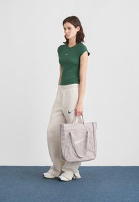 Νεαρή γυναίκα φοράει πράσινο εφαρμοστό πουκάμισο και μπεζ φούτερ, στέκεται σε μπλε χαλί κρατώντας μια μεγάλη ανοιχτό γκρι τσάντα ώμου με λευκό λογότυπο.