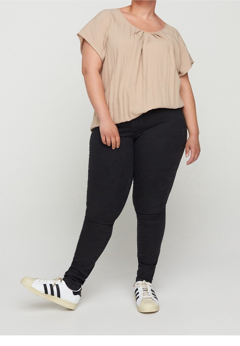 Personne portant un chemisier beige à manches courtes, un pantalon noir ajusté et des baskets blanches avec des rayures noires, debout avec une jambe tendue vers l'avant.