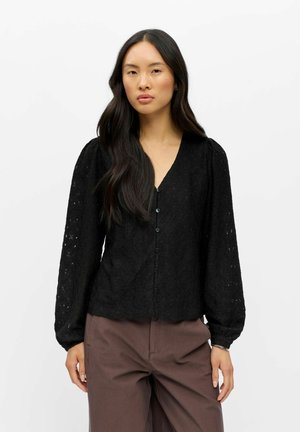 Femme aux longs cheveux foncés portant un chemisier noir texturé à boutons avec des manches bouffantes et un pantalon marron, debout sur un fond uni.