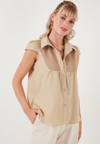 Beige bezramenný top s krajkovými detaily na ramenou, zapínání na knoflíky vepředu, měkká textura a zakřivený lem.