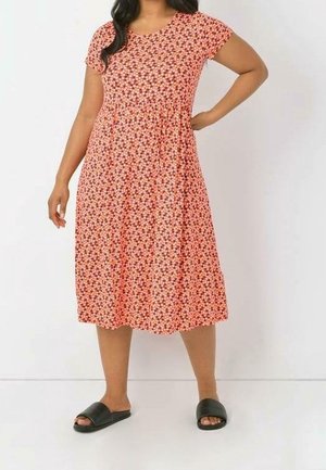 Nainen pukeutuneena polvipituiseen, lyhythihaiseen oranssiin mekkoon, jossa pieni kukkakuosi, sekä mustiin slip-on-sandaaleihin, seisoo yksiväristä seinää vasten.