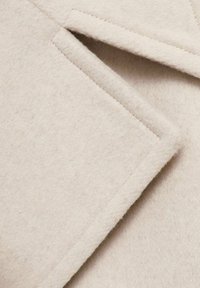 Beżowa wełniana tkanina o miękkiej, teksturowanej powierzchni, z ostrym kołnierzem i ukośnymi klapami, starannie wykończonymi brzegami.