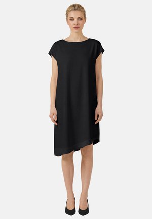 Blonde Frau steht und trägt ein einfaches schwarzes Kleid mit kurzen Ärmeln, einem asymmetrischen Saum und schwarzen Absätzen vor einem schlichten grauen Hintergrund.