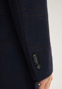 Nahaufnahme eines navyblauen Wollblazers mit strukturiertem Stoff, dezenten braunen Streifen und vier schwarzen Knöpfen am Ärmelbund.