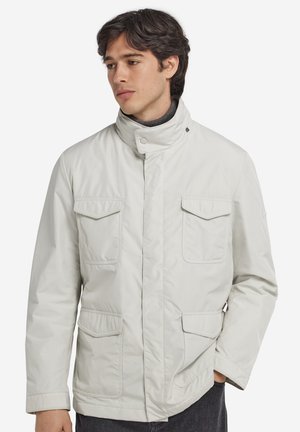 Ung mand iført en lys beige jakke med fire klaplommer og en høj krave, der kigger let til siden mod en ensfarvet baggrund.