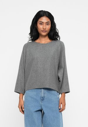 Hallhõlmade, pehmest kangast, lahti istuv hall kampsun ümmarguse kaelusega ja laiad varrukad, millele on kombineeritud kõrge vöökohaga heledad jeans.