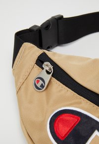Banane en tissu beige avec une sangle noire, dotée d'une petite tirette en métal argenté et d'un logo multicolore bien visible. Surface texturée.