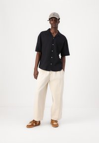 Camisa negra de manga corta y botones con un bolsillo en el pecho, combinada con pantalones anchos color crema. Zapatillas marrones con acentos texturizados.