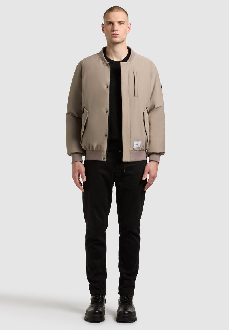 Barna bomber dzseki sima felülettel, bordás mandzsettákkal, cipzáras zsebekkel és logóval az alján, fekete nadrággal és cipővel párosítva.