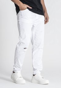 Vaqueros blancos desgastados con rasgaduras visibles, de algodón, diseño de corte recto. Combinados con zapatillas blancas sobre un fondo neutro.