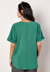 Maglietta verde a maniche corte con vestibilità morbida, scollatura rotonda e risvolti. Il tessuto testurizzato ha una leggera lucentezza e un orlo curvo.
