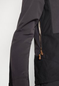Svart ytterjakke med glidelåslomme og beige detaljer, glatt tekstur og tettsittende ermedesign, som fremhever holdbare materialer.
