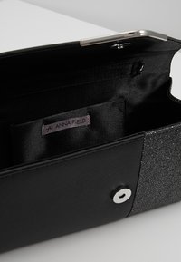 Černé psaníčko s texturovaným povrchem, stříbrnými kovovými doplňky a třpytivým akcentem. Uvnitř je kapsa s nášivkou "ANNA FIELD".