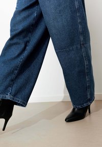 Jean denim coupe large en bleu foncé, présentant une coupe droite et des coutures apparentes. Associé à des talons noirs pointus, mettant en avant un design élégant.