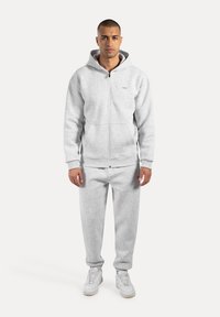 Grauer Zip-Hoodie mit Fronttasche, kombiniert mit passenden grauen Jogginghosen. Sanfte Textur, lässige Passform und weiße Sneakers.