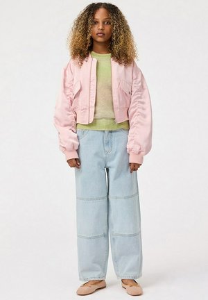 Giacca bomber in satin rosa, top lavorato a maglia verde chiaro, jeans oversize azzurri con cuciture e ballerine beige. Capelli ricci e sfondo neutro.
