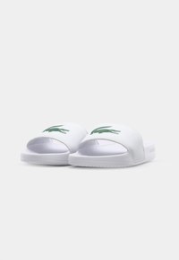 Sandales blanches à semelle plate avec un logo crocodile vert, fabriquées en matériau synthétique, dotées d'un design à bout ouvert et d'une semelle texturée.