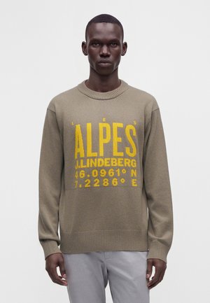 Un homme porte un pull en maille beige avec un grand texte jaune ""ALPES J. LINDEBERG"" et des coordonnées, associé à un pantalon gris clair, debout devant un fond blanc.