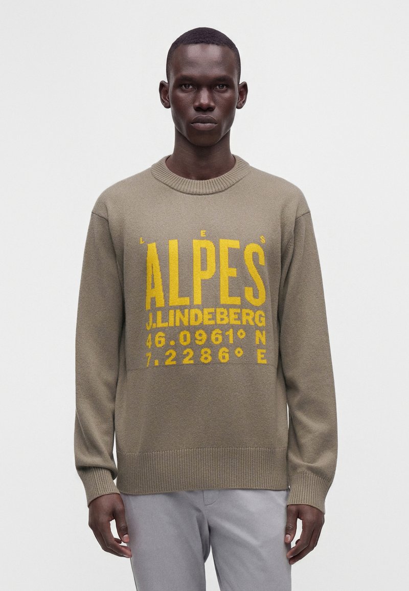 Un homme porte un pull en maille beige avec un grand texte jaune ""ALPES J. LINDEBERG"" et des coordonnées, associé à un pantalon gris clair, debout devant un fond blanc.
