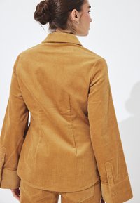 Ženska z rjavimi lasmi v figi, oblečena v blazer iz manjših žametnih črt v gorčično rumeni barvi in ujemajoče se hlače, obrnjena z zadnje proti enostavni beli podlagi.