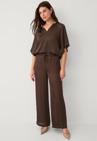 Brun, let jumpsuit med V-udskæring og brede ærmer; har en elastisk talje og løse brede ben. Båret med beige hæle.