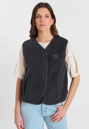 Vrouw draagt een zwart fleecevest over een wit kortärmelig shirt en lichtblauwe spijkerbroek, staand tegen een witte achtergrond.