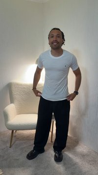 Man standing indoors wearing a white t-shirt and black pants with black shoes near a beige textured armchair and lamp.
