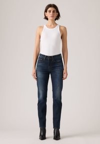 Γυναίκα που στέκεται, φορώντας λευκή αμάνικη μπλούζα, σκούρο μπλε τζιν και μαύρα μποτάκια αστραγάλου, μπροστά σε απλό ανοιχτό φόντο.