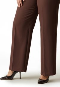 Pantaloni marroni a gamba larga realizzati in tessuto liscio, abbinati a décolleté nere lucide con punta appuntita e una texture sottile.