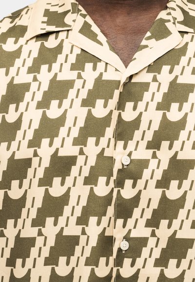 Κουμπωτό πουκάμισο με κοντά μανίκια και γεωμετρικό μοτίβο σε ελαιόπρασινο και μπεζ χρώμα στην περιοχή του άνω στήθους και της κλείδας ενός ατόμου.