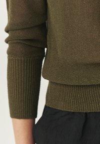 Olíva zöld kötött pulóver, bordás aljjal és mandzsettákkal, texturált mintázattal és enyhén áttetsző ujjakkal.
