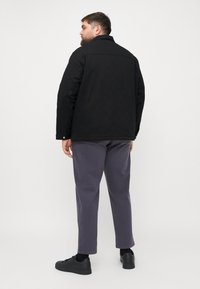 Zwarte jas met een kraag en knoopmanchetten, gecombineerd met grijze broek en zwarte schoenen. De stof lijkt soepel met een losse pasvorm.