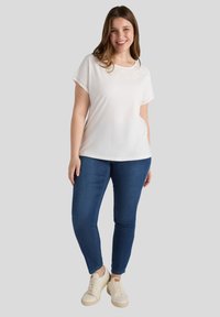 Camiseta blanca de manga corta con detalles de ojales decorativos en el escote, combinada con vaqueros pitillo azules y zapatillas beige.