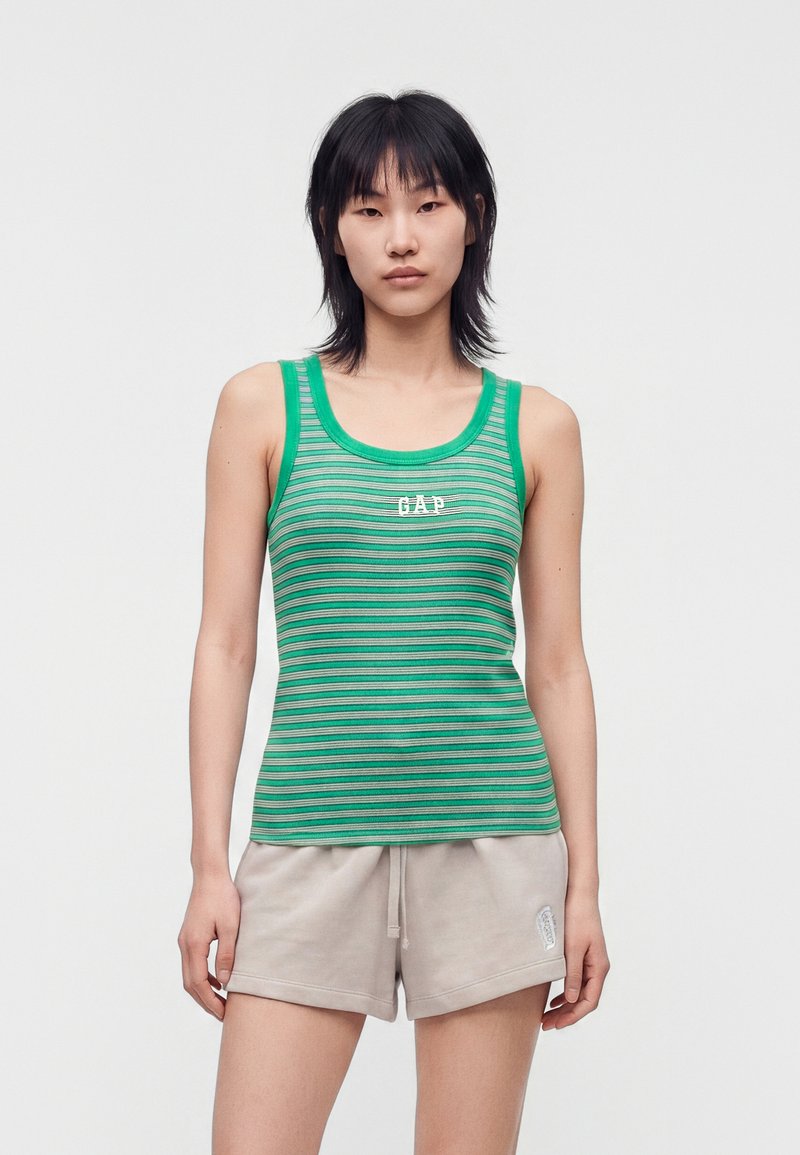 Femme debout devant un fond uni, portant un débardeur vert à rayures avec le texte "GAP" et un short beige à cordon, face à l’avant.