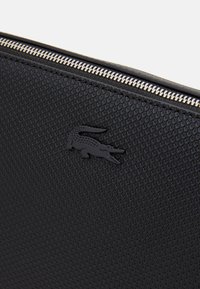 Schwarze, strukturierte Tasche mit einem glatten Reißverschluss und einem erhabenen schwarzen Krokodil-Logo. Das Material weist ein netzartiges Muster auf.