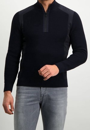 Homme portant un pull noir à fermeture éclair mi-longue avec un tricot côtelé et des empiècements solides aux épaules, associé à un jean gris clair, debout devant un fond uni.