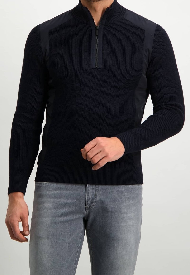 Homme portant un pull noir à fermeture éclair mi-longue avec un tricot côtelé et des empiècements solides aux épaules, associé à un jean gris clair, debout devant un fond uni.