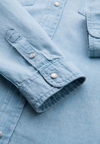 Manche de chemise en denim bleu clair avec poignet boutonné, présentant des boutons-pression argentés et des coutures visibles, montrant une texture douce.