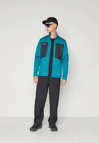 Man i tealjacka med svarta bröstfickor, svarta byxor, svart keps, reflexiv solglasögon och svarta vandringsskor, står och tittar åt vänster.