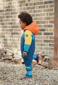 Kleurvolle waterdichte overall in blauw, geel en oranje, met een kap, een ritszak en blauwe rubberen laarzen met oranje zolen.