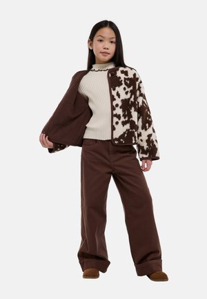 Jeune fille portant un pantalon large marron, un pull côtelé crème et une veste marron et crème à motifs, debout avec un côté ouvert.