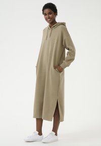 Hooded, lange beige jurk met zijsplitten, heeft kangoeroe zakken en geborduurd logo. Gewaardeerd met witte sneakers op een neutrale achtergrond.