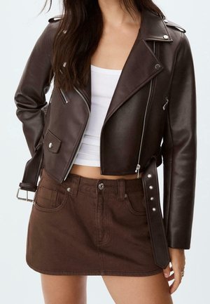 Vrouw die een donkerbruine leren motorjack draagt met zilveren ritsen en riem, een wit cropped tanktop en een donkerbruine denim minirok.