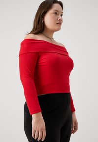 Piros, vállnál kivágott, bordázott felső hosszú ujjakkal. Az anyag sima és rugalmas, fekete, feszes leggingsszel párosítva.